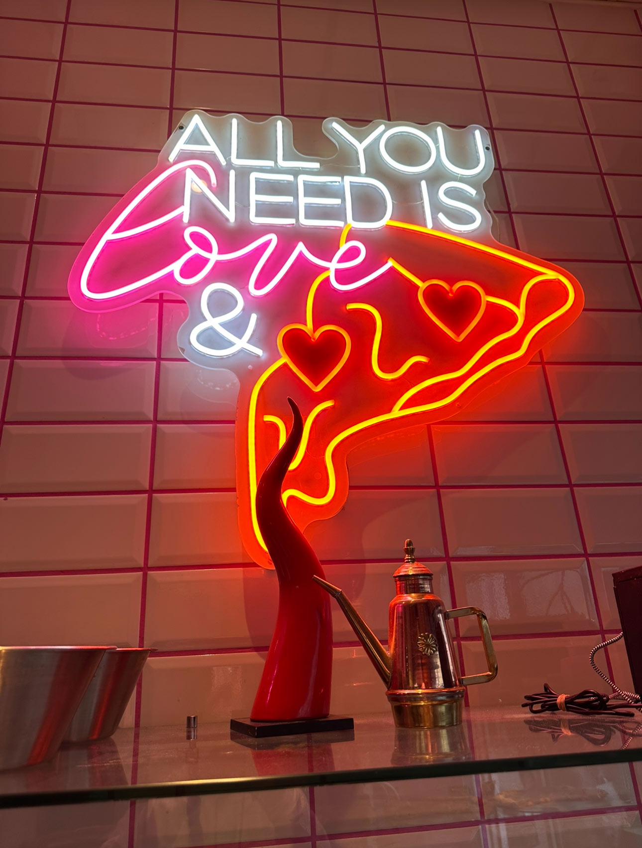 Neon-Leuchtschild mit "All you need is love" und rote Pizza mit Herzen auf rosa Fliesenwand.
